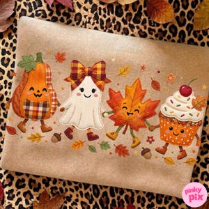 May include: A tan fabric surface featuring an embroidered design of four cartoon characters: an orange pumpkin, a white ghost with a plaid bow, an orange maple leaf, and a cupcake, all surrounded by small scattered autumn leaves and acorns.