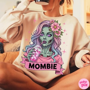 May include: Beige sweatshirt featuring a zombie graphic with pink and purple hair, holding a pink drink. The graphic includes the word "MOMBIE" on a pink banner, with pink star accents.