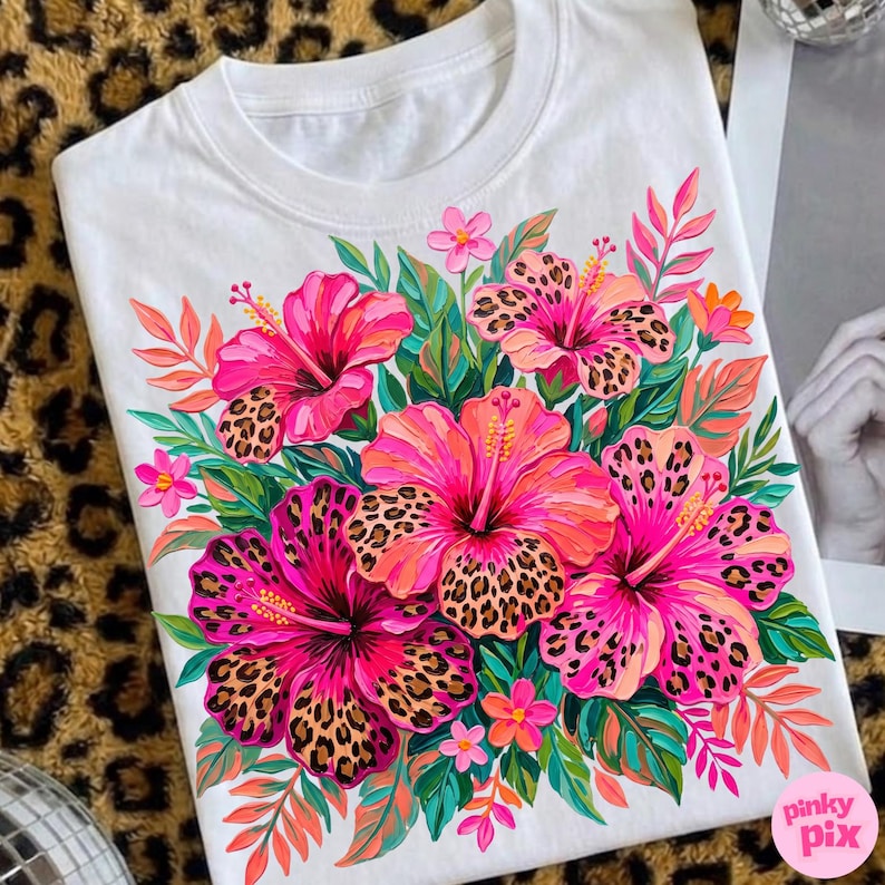 May include: White t-shirt featuring a vibrant floral design. The design showcases large pink hibiscus flowers with leopard print centers, surrounded by green leaves and smaller pink flowers. The overall aesthetic is bright and tropical.