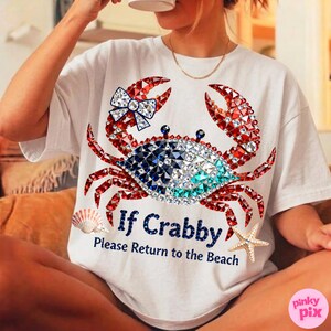 May include: White t-shirt featuring a crab design with red, blue, and clear crystal accents. The crab has a bow on one claw. Text on the shirt reads "If Crabby, Please Return to the Beach". A seashell and starfish are also on the shirt.