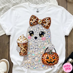 Może przedstawiać: Biały t-shirt z grafiką ducha ozdobionego przezroczystymi, opalizującymi cyrkoniami i perłami. Duch ma na sobie kokardę w panterkę, trzyma kubek do kawy w panterkę i niesie pomarańczowe wiaderko w kształcie dyni na Halloween.