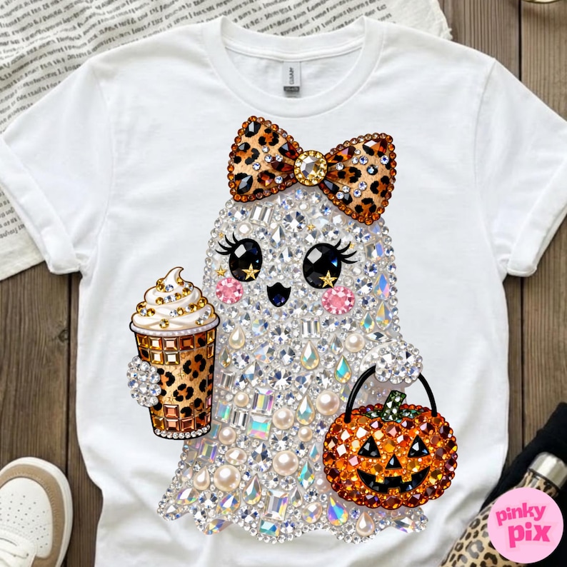 Może przedstawiać: Biały t-shirt z grafiką ducha ozdobionego przezroczystymi, opalizującymi cyrkoniami i perłami. Duch ma na sobie kokardę w panterkę, trzyma kubek do kawy w panterkę i niesie pomarańczowe wiaderko w kształcie dyni na Halloween.