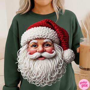 May include: A dark green sweatshirt featuring a detailed Santa Claus face with a red and white knitted hat and long white beard. The Santa face is the focal point, with blue eyes and a rosy complexion. The sweatshirt is a solid color.
