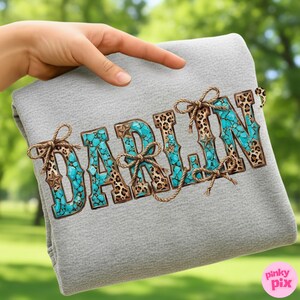 Pu&ograve; includere: Una coperta grigia con la scritta "DARLIN" in turchese, stampa leopardata e dettagli in corda marrone. Le lettere sono riempite di pietre turchesi e stampa leopardata, con dettagli a stella. La coperta &egrave; tenuta da una mano.