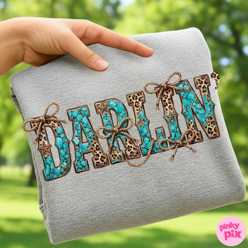 Pu&ograve; includere: Una coperta grigia con la scritta "DARLIN" in turchese, stampa leopardata e dettagli in corda marrone. Le lettere sono riempite di pietre turchesi e stampa leopardata, con dettagli a stella. La coperta &egrave; tenuta da una mano.