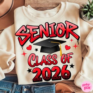 May include: Cream-colored sweatshirt with the words "SENIOR CLASS OF 2026" in red graffiti-style lettering. A black graduation cap with a gold tassel is centered above the words. Red hearts and stars are scattered around the text.