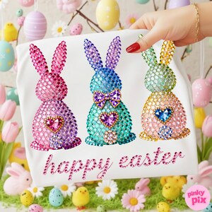 May include: A white Easter-themed decorative cloth featuring three jeweled bunny figures in pink, blue, and yellow. Each bunny has a heart-shaped gem. The words "happy easter" are written in pink script. The background includes Easter eggs and flowers.