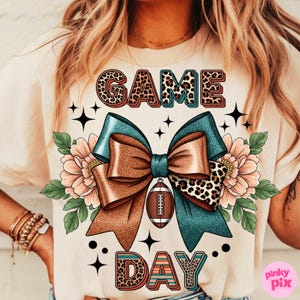 May include: Cream-colored t-shirt with a "GAME DAY" design. The design features a large bow with teal and brown ribbons, a football, and floral accents. The words "GAME" and "DAY" are in a decorative font with leopard print.