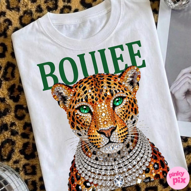 May include: White t-shirt featuring a jeweled leopard design with green eyes and a diamond necklace. The word "BOUIEE" is printed in green above the leopard. The shirt is on a leopard print background.