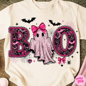 Puede incluir: Camiseta color crema con un dise&ntilde;o tem&aacute;tico de Halloween. La palabra "BOO" est&aacute; escrita en letras rosas con detalles de encaje negro. Un fantasma rosa con un lazo est&aacute; en el centro, rodeado de murci&eacute;lagos, telara&ntilde;as y estrellas.