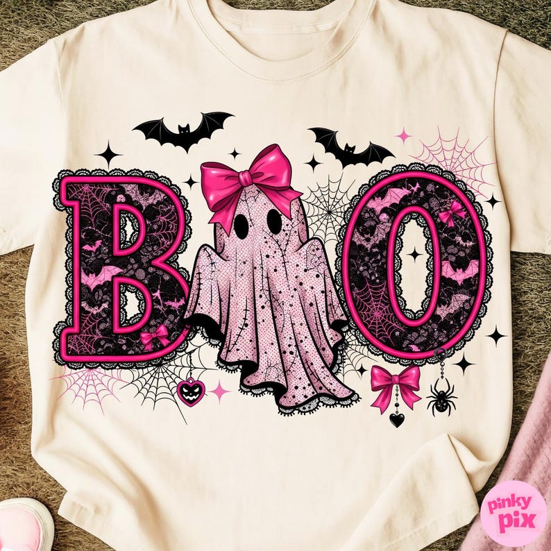 Puede incluir: Camiseta color crema con un dise&ntilde;o tem&aacute;tico de Halloween. La palabra "BOO" est&aacute; escrita en letras rosas con detalles de encaje negro. Un fantasma rosa con un lazo est&aacute; en el centro, rodeado de murci&eacute;lagos, telara&ntilde;as y estrellas.