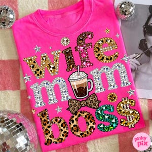 Puede incluir: Una camiseta rosa brillante con las palabras "Wife Mom Boss" en pedrería y estampado de leopardo. También incluye una taza de café, una pajarita y un signo de dólar. La camiseta está decorada con pedrería en forma de estrella.