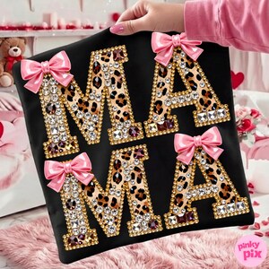 May include: Black t-shirt featuring the word "MAMA" in leopard print letters, adorned with rhinestones and gold edging. Each letter is decorated with a pink bow. The shirt is held up by a person.