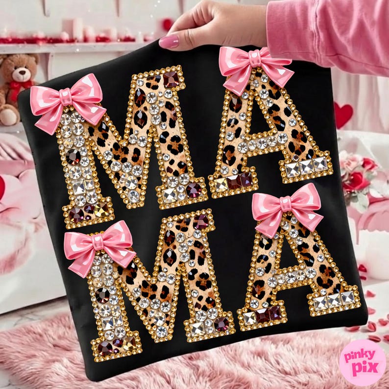 May include: Black t-shirt featuring the word "MAMA" in leopard print letters, adorned with rhinestones and gold edging. Each letter is decorated with a pink bow. The shirt is held up by a person.