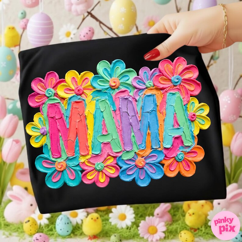May include: Black t-shirt with the word "MAMA" in colorful, textured letters, surrounded by vibrant, multi-colored flowers. The design features a palette of pink, blue, yellow, and orange. The shirt is held up by a hand with red nail polish.
