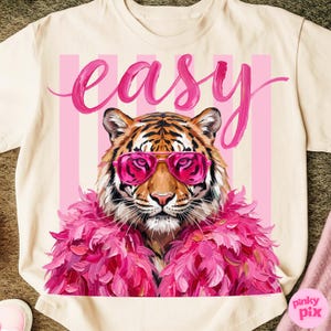 May include: Cream-colored t-shirt featuring a tiger wearing pink sunglasses and a pink feather boa. The word "easy" is written in pink script above the tiger. Pink vertical stripes are behind the tiger.