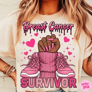 May include: Cream-colored t-shirt with a pink graphic design. The design includes the words "Breast Cancer Survivor" in pink, a raised fist, pink hearts, and pink sneakers.