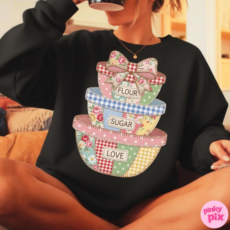 May include: Black sweatshirt featuring a stack of patchwork-style mixing bowls. The bowls are decorated with floral, gingham, and polka dot patterns. Text on the bowls reads "FLOUR", "SUGAR", and "LOVE". A large bow tops the stack.