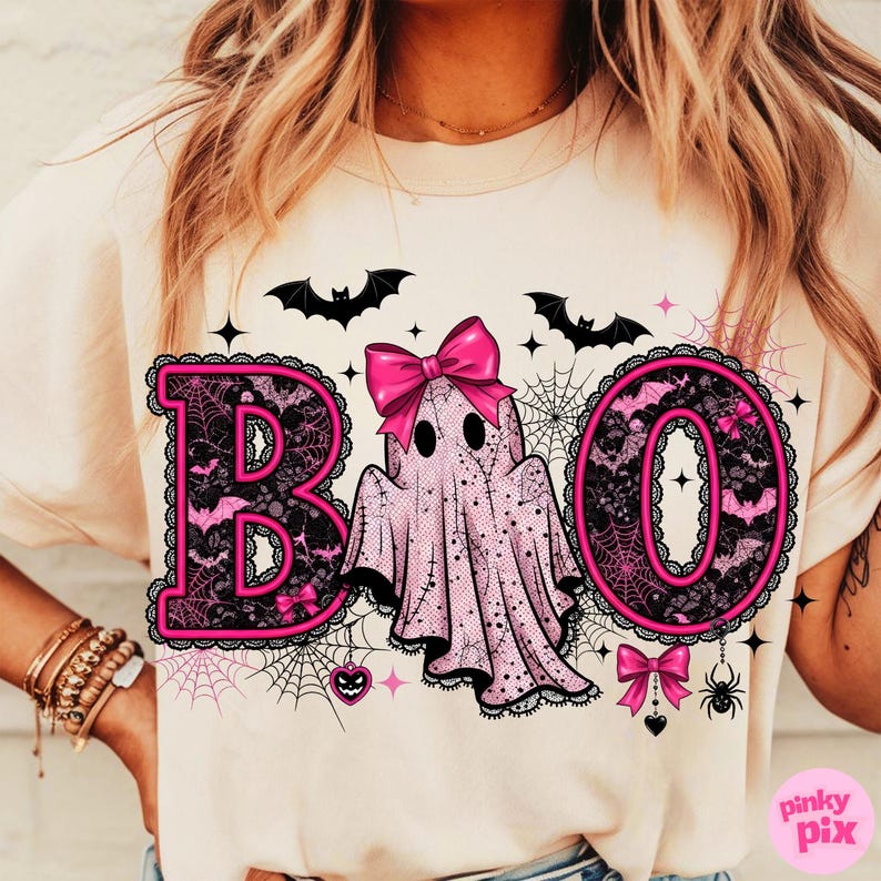 Puede incluir: Camiseta de color beige claro con un dise&ntilde;o tem&aacute;tico de Halloween. La palabra "BOO" se muestra en rosa, con un fantasma en el centro. El dise&ntilde;o incluye murci&eacute;lagos, telara&ntilde;as y lazos, creando una est&eacute;tica festiva y espeluznante.