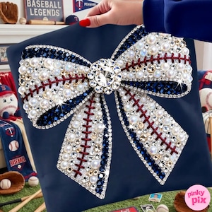 May include: A navy blue pillow with a large bow design. The bow is embellished with pearls, rhinestones, and dark blue accents, mimicking a baseball. Red stitching detail adds to the baseball theme. A large, sparkling crystal sits in the center.