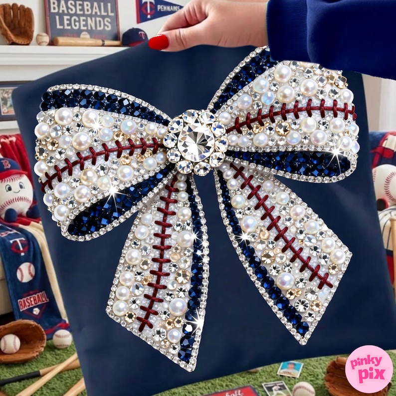 May include: A navy blue pillow with a large bow design. The bow is embellished with pearls, rhinestones, and dark blue accents, mimicking a baseball. Red stitching detail adds to the baseball theme. A large, sparkling crystal sits in the center.