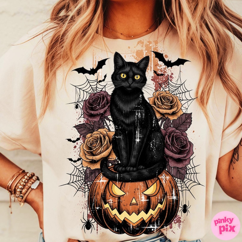 May include: Cream-colored t-shirt featuring a black cat with yellow eyes sitting on a carved pumpkin. The design includes purple and gold roses, spiderwebs, bats, and spiders, creating a Halloween theme.