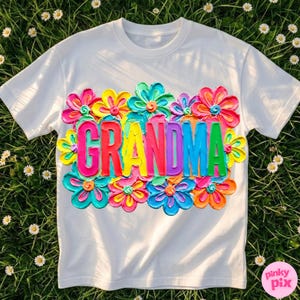 May include: White t-shirt with the word "GRANDMA" in colorful, textured letters, surrounded by vibrant, multi-colored flower designs. The shirt is made of a soft material and is perfect for a gift.