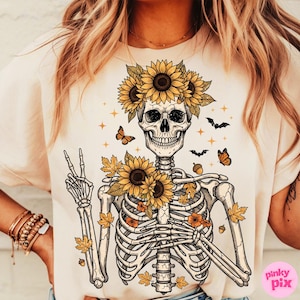 May include: Cream-colored t-shirt featuring a skeleton design adorned with sunflowers, autumn leaves, and butterflies. The skeleton is wearing a sunflower crown and giving a peace sign. The design includes bats and acorns.