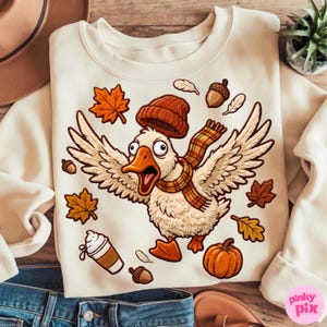 May include: A cream-coloured sweatshirt featuring a graphic of a white goose wearing a brown knit beanie and a plaid scarf. The goose is surrounded by orange maple leaves, brown acorns, a small pumpkin, and a cup of coffee with whipped cream.