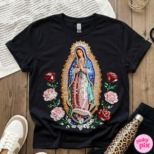 May include: Black t-shirt featuring a detailed graphic of the Virgin Mary with a mosaic-like design, surrounded by red and pink roses. The design includes blue, red, and gold colors. The shirt is displayed on a wooden surface.