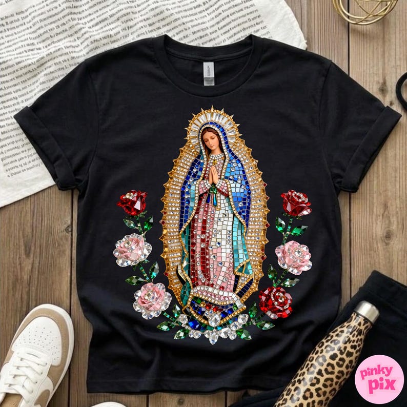 May include: Black t-shirt featuring a detailed graphic of the Virgin Mary with a mosaic-like design, surrounded by red and pink roses. The design includes blue, red, and gold colors. The shirt is displayed on a wooden surface.