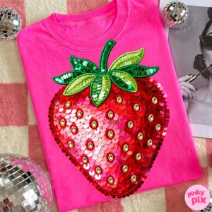 Pode incluir: Uma camisola rosa brilhante com um grande desenho de morango com lantejoulas. O morango é vermelho com detalhes dourados e folhas verdes. A camisola é de gola redonda.