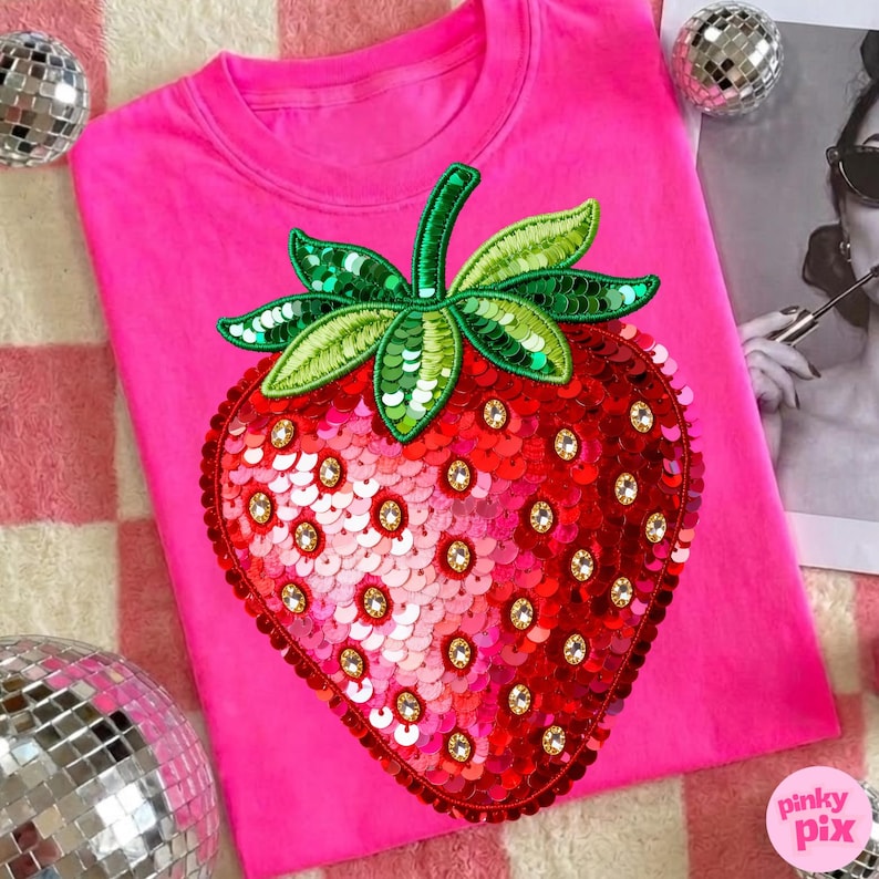 K&ouml;nnte beinhalten: Ein leuchtend pinkfarbenes Sweatshirt mit einem gro&szlig;en, paillettenbesetzten Erdbeermotiv. Die Erdbeere ist rot mit goldenen Akzenten und gr&uuml;nen Bl&auml;ttern. Das Shirt hat einen Rundhalsausschnitt.