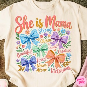 K&ouml;nnte beinhalten: Cremefarbenes T-Shirt mit den Worten "She is Mama" in rosa und gelber Schrift. Das Shirt zeigt bunte Schleifen und florale Akzente mit W&ouml;rtern wie "Strong", "Beautiful" und "Victorious".