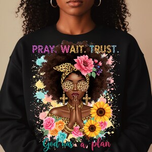 May include: Black sweatshirt with a colorful graphic design featuring a woman with a leopard print headband and sunglasses, praying with her hands clasped. The design includes flowers, sunflowers, and the text "Pray. Wait. Trust. God has a plan."