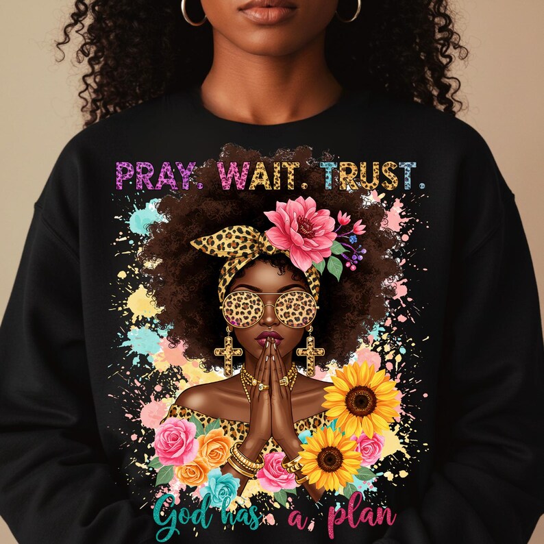 May include: Black sweatshirt with a colorful graphic design featuring a woman with a leopard print headband and sunglasses, praying with her hands clasped. The design includes flowers, sunflowers, and the text "Pray. Wait. Trust. God has a plan."