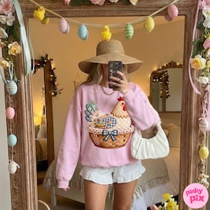 May include: A light pink sweatshirt with a chicken design in a basket. The image is reflected in a mirror decorated with Easter eggs and flowers. The person in the image is wearing a straw hat and white shorts.