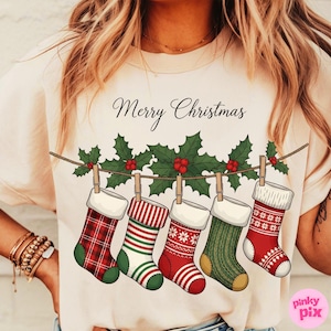 May include: A cream-colored t-shirt featuring a festive design. The shirt displays five Christmas stockings hanging from a string, each with a unique pattern and color. Above the stockings is the text "Merry Christmas".