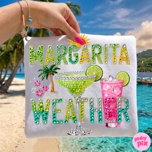 Peut inclure: Serviette de plage blanche avec l'inscription "MARGARITA WEATHER" en lettres vertes et dor&eacute;es scintillantes. La serviette pr&eacute;sente un verre &agrave; margarita, un cocktail rose, un palmier et des tranches de citron vert, le tout orn&eacute; de strass. Une journ&eacute;e ensoleill&eacute;e &agrave; la plage.