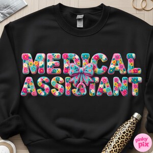 May include: Black sweatshirt with the words "MEDICAL ASSISTANT" in large, colorful, floral letters. A blue bow and stethoscope graphic are centered between the words. The sweatshirt is a crew neck style.