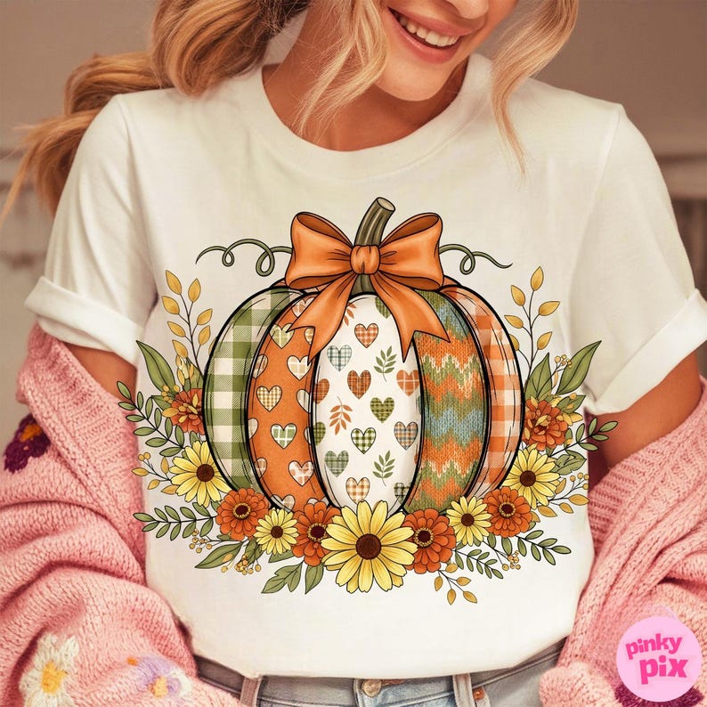 May include: White t-shirt featuring a colorful pumpkin design. The pumpkin has sections with plaid, hearts, and a knit pattern, topped with an orange bow. Floral accents include sunflowers and leaves, perfect for fall.