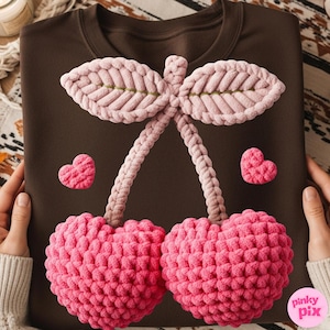 May include: A brown sweatshirt featuring a large, pink, crocheted cherry design with two hearts. The cherry has a textured appearance, with a light pink stem and leaves. The design is centered on the front of the shirt.