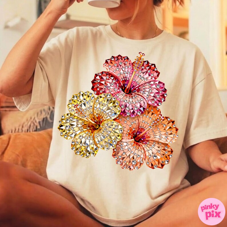 May include: Cream-colored t-shirt featuring a vibrant floral design. The design showcases three hibiscus flowers in shades of red, pink, orange, and yellow, with a sparkling, jeweled effect. The shirt is a relaxed fit.