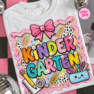 May include: White t-shirt with a colorful "Kindergarten" graphic. The design features a pink bow, a decorative border, and school supplies like pencils and scissors. The text is in bright colors with flower accents.