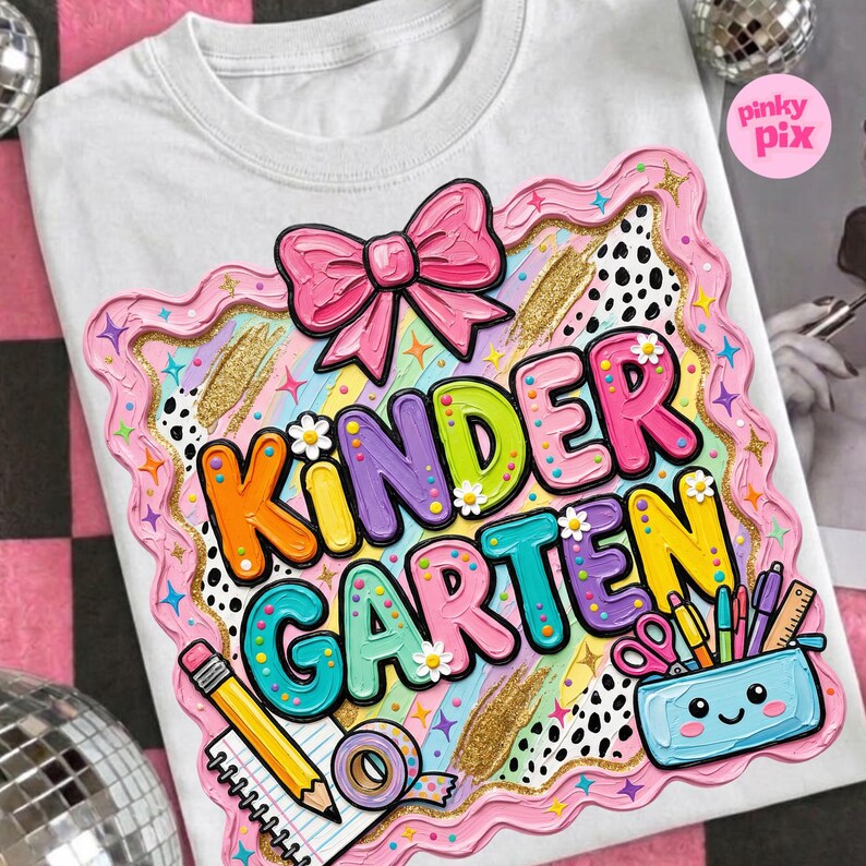 May include: White t-shirt with a colorful "Kindergarten" graphic. The design features a pink bow, a decorative border, and school supplies like pencils and scissors. The text is in bright colors with flower accents.
