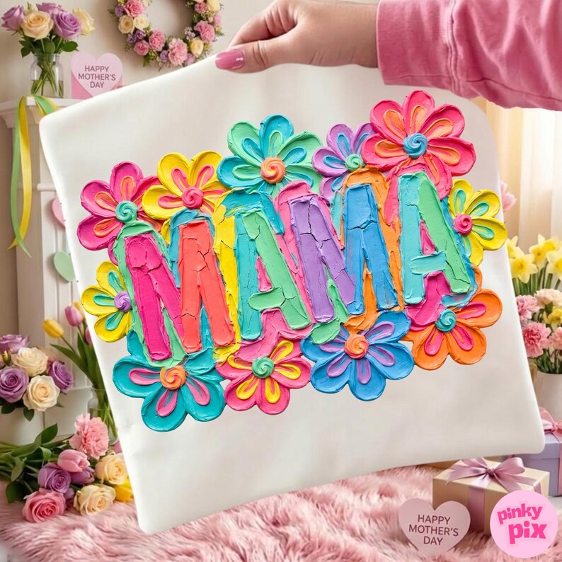 May include: A white pillow with the word "MAMA" in colorful, textured paint, surrounded by vibrant, multi-colored flowers. The pillow is held up by a person. The theme is Mother's Day.