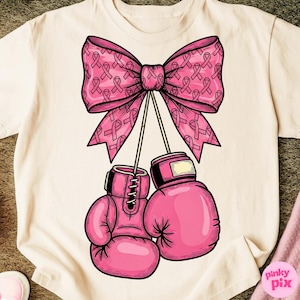 May include: Cream-colored t-shirt featuring a pink bow adorned with breast cancer awareness ribbons. Below the bow, two pink boxing gloves hang, symbolizing strength and support. The design is a graphic representation of solidarity.