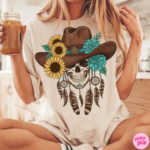 Op de afbeelding: Beige T-shirt met een schedelontwerp met een bruine cowboyhoed versierd met zonnebloemen, turquoise bloemen en een ster. Dromenvangers en veren hangen eronder. Het shirt heeft een losse pasvorm.