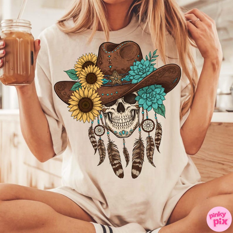 Op de afbeelding: Beige T-shirt met een schedelontwerp met een bruine cowboyhoed versierd met zonnebloemen, turquoise bloemen en een ster. Dromenvangers en veren hangen eronder. Het shirt heeft een losse pasvorm.