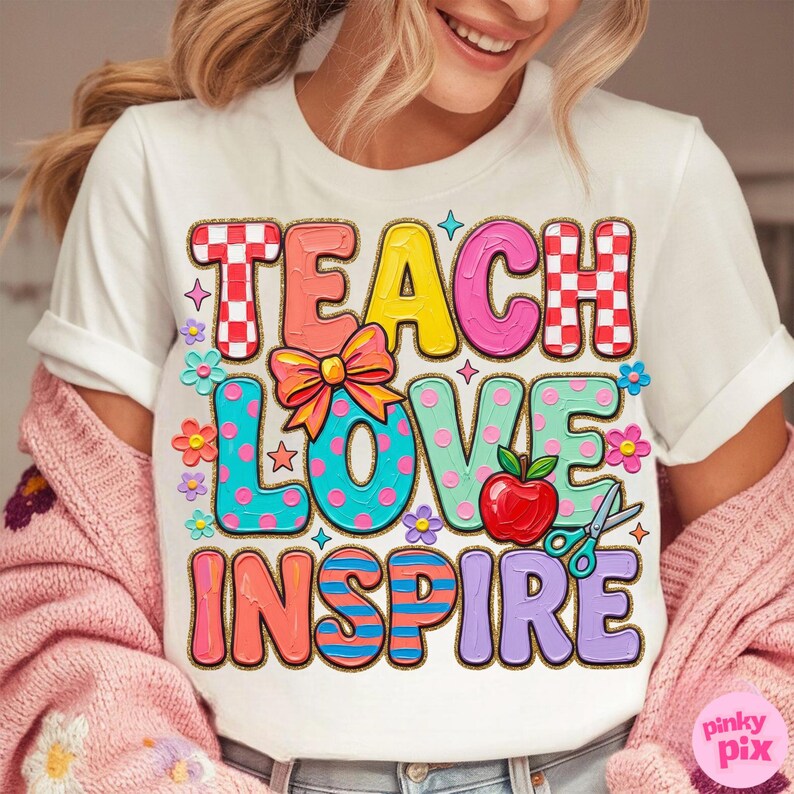 May include: White t-shirt with the words "TEACH LOVE INSPIRE" in colorful, playful lettering. The design includes a red apple, scissors, and floral accents. The shirt is a casual wear item.